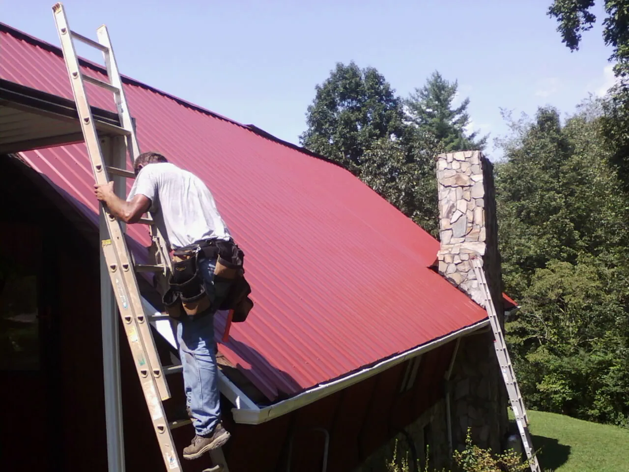 Roofer on a ladder beside a red metal roof and stone chimney — Soffit & Fascia Repair by Roofing Brothers in Currie