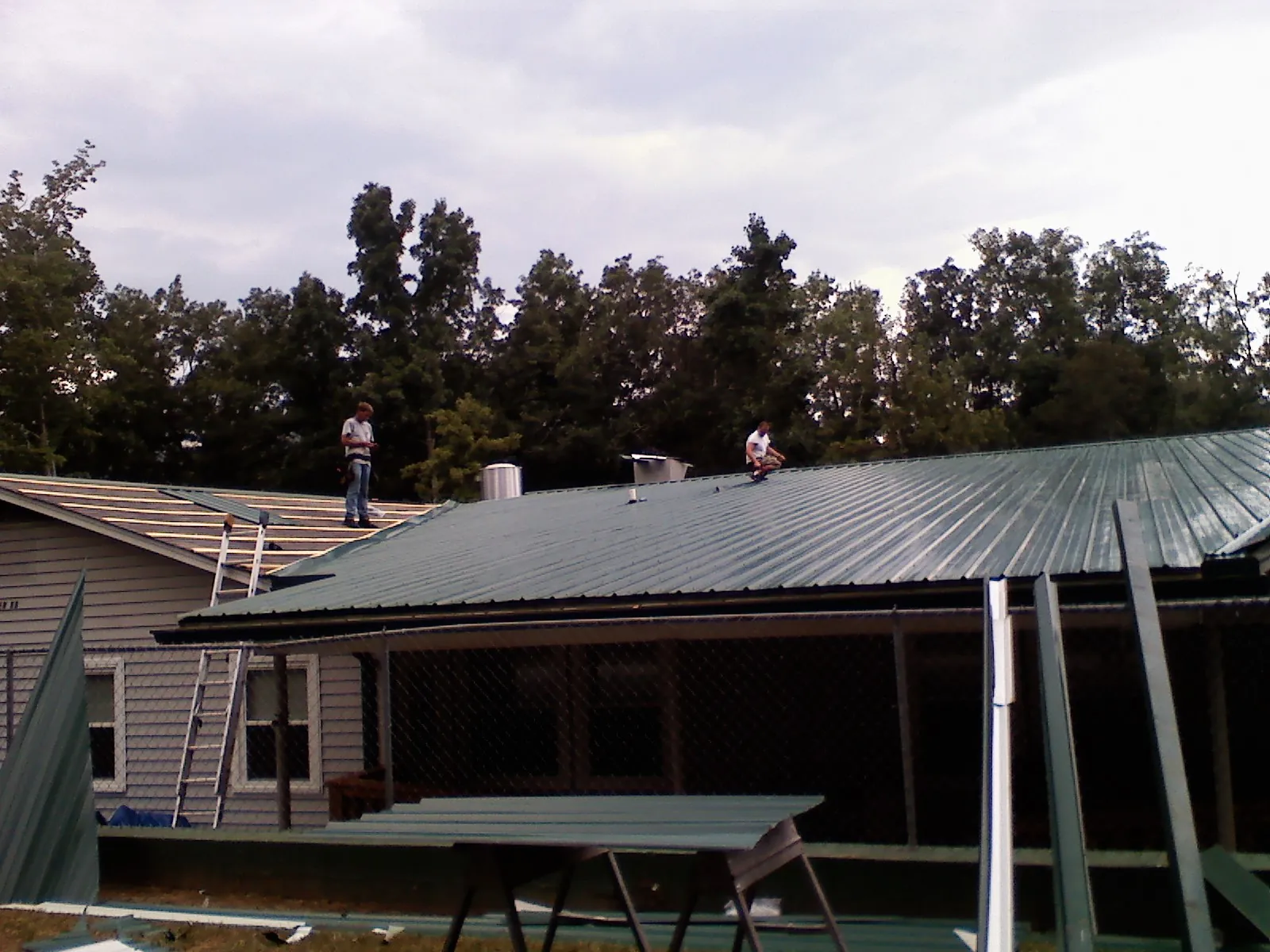 Two roofers installing green metal panels above a covered porch in Currie
